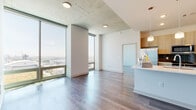 This single-floor high-rise unit offers a refined urban living experience defined by clean lines, modern finishes, and an abundance of natural light. The open-concept living and dining area is anchored by dramatic floor-to-ceiling glass walls that frame sweeping panoramic city views, creating a bright and airy atmosphere throughout. Exposed concrete ceiling panels add an industrial-modern aesthetic contrast to the otherwise crisp white walls.

The kitchen is thoughtfully designed with light wood cabinetry, stainless steel appliances including a refrigerator, range, microwave, and dishwasher, as well as a teal glass tile backsplash that adds a subtle pop of color. A generous island with a white countertop and undermount sink serves as both a functional workspace and casual gathering point, complemented by pendant lighting overhead.

Hardwood flooring flows consistently throughout the living areas and bedrooms, lending warmth and continuity to the space. The bedrooms feature large floor-to-ceiling windows and sliding glass doors that open to outdoor access, each offering unobstructed elevated views of the cityscape below.

The bathrooms are finished with marble-style tile on both the floors and walls. One bathroom features a soaking tub with a shower combination, while the second offers a sleek glass-enclosed walk-in shower. A hallway with louvered and paneled doors provides access to closets and additional rooms, completing this well-appointed urban residence.