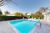 Located at 901 Lincoln Rd in Yuba City, California, this outdoor community pool area offers a well-appointed recreational space designed for relaxation and leisure. The centerpiece is a generously sized rectangular swimming pool with crystal-clear turquoise water, accessible via stainless steel handrails on multiple sides. The pool is flanked by a wide concrete deck that provides ample space for lounging, with white patio chairs, lounge chairs, and a shaded umbrella conveniently arranged poolside. The perimeter of the pool area is enclosed by a tall, manicured hedge that creates a sense of privacy and seclusion while framing views of mature trees, including a prominent deciduous tree that adds natural character to the setting. A dedicated pool house structure is visible on one side, featuring a gated entry and informational signage, suggesting organized management of the amenity. The surrounding buildings display green awnings and neutral siding, contributing to a cohesive and tidy aesthetic. Under the open California sky, the space benefits from abundant natural light, making it an inviting outdoor retreat. The combination of practical amenities, thoughtful landscaping, and a well-maintained pool makes this a standout communal feature of the property.