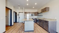 This single-floor unit offers a thoughtfully designed layout with modern finishes throughout. The kitchen serves as a central highlight, showcasing dark-stained cabinetry with sleek hardware, light granite countertops, and a full suite of stainless steel appliances including a refrigerator, range, over-the-range microwave, and dishwasher. A center island provides additional counter space and storage, while light wood-style flooring ties the area together, creating a warm and functional atmosphere. Recessed lighting illuminates the space efficiently.

The bedroom areas feature neutral carpet flooring and large windows trimmed in dark accents, allowing natural light to fill the rooms while offering views of the surrounding neighborhood. Baseboard heating units are present beneath the windows, ensuring comfort year-round.

The bathroom continues the cohesive design aesthetic with matching dark cabinetry, granite countertops, an undermount sink, and a large mirror above the vanity. Wood-look tile flooring grounds the space with a clean, contemporary feel.

Overall, the unit presents a consistent, modern design language with quality finishes, practical storage solutions, and well-proportioned rooms that balance everyday functionality with visual appeal.