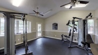 This fitness center is thoughtfully designed to support a comprehensive workout experience across multiple connected zones. The main workout area features vaulted ceilings with dual ceiling fans for air circulation, neutral gray walls with a classic white chair rail, and durable dark rubber flooring throughout. Natural light filters in through multiple windows, offering views of the surrounding greenery, while a set of French doors provides convenient access to the outdoors. Strength training equipment, including a power rack and a multi-functional cable machine with a dedicated bench, anchors this primary space. A posted fitness center rules board keeps the environment organized and community-oriented. An adjacent cardio zone is outfitted with a treadmill, elliptical trainer, upright stationary bike, and a rowing machine, all positioned in front of a large wall mirror that enhances the sense of space. A wall-mounted flat-screen television adds entertainment to cardio sessions. A separate alcove area connects to an additional room, visible through a windowed interior door, and includes practical amenities such as sanitation dispensers, a fire extinguisher, and waste receptacles, ensuring the facility is safe and well-maintained throughout.