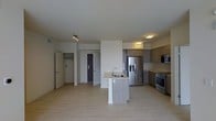 This single-floor space showcases a contemporary open-concept layout that seamlessly integrates the kitchen and living areas. The kitchen is a standout feature, centered around a freestanding island with a built-in sink and light wood-toned cabinetry that extends to the perimeter walls. Upper and lower cabinets in a warm taupe-gray finish provide generous storage, while stainless steel appliances — including a French door refrigerator with water and ice dispenser, a range, and an over-the-range microwave — add a sleek, modern touch. Light hardwood-style flooring runs throughout the space, creating a bright and cohesive aesthetic that complements the white walls and ceilings. Flush-mounted and track lighting fixtures provide consistent illumination across the open area. In the background, multiple interior doors are visible — one leading to what appears to be a closet with a rod and shelving, and another suggesting access to a separate room. The overall ambiance is clean, minimalist, and functional, with a neutral palette that lends itself to a variety of interior design styles.