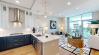 This two-bedroom residence at the Emerson in Tampa, FL offers a well-appointed single-floor layout with contemporary design elements and high-end finishes. The open-concept living and kitchen area is immediately striking, featuring light hardwood flooring, recessed lighting, and floor-to-ceiling windows that flood the space with natural light and provide sweeping outdoor views of the Tampa skyline and surrounding greenery.

The kitchen is a standout, showcasing a two-tone design with navy lower cabinetry paired with crisp white upper cabinets, all accented by gold hardware. A large quartz waterfall island, stainless steel appliances, a gas cooktop, built-in wall oven, and a textured tile backsplash complete the space. Gold cage pendant lights add a refined touch overhead.

The living area flows seamlessly from the kitchen, offering an inviting, light-filled atmosphere with views to the outdoors. A built-in bar area with a wine cooler adds a functional and stylish element nearby.

Both bedrooms are generously sized, with large windows bringing in abundant natural light. The primary bedroom connects to an en-suite bathroom featuring a navy vanity with gold accents, white quartz countertops, and a tiled walk-in shower. A spacious walk-in closet with ample shelving and hanging storage serves the primary suite. Additional bathroom finishes mirror the same polished aesthetic found throughout the residence.