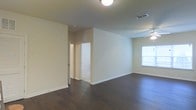 This single-story space presents a straightforward and functional layout, characterized by its clean, neutral aesthetic. The main living area features dark hardwood-style flooring that contrasts with the light beige walls, creating a grounded, cohesive look throughout. A ceiling fan is mounted overhead, providing both practical air circulation and a modest focal point to the room.

Natural light filters in through a window positioned along one wall, lending the space a bright and airy feel. Overhead lighting fixtures supplement illumination in areas less reached by natural light. The walls are smooth and painted in a warm neutral tone, offering a versatile backdrop suitable for various interior styles.

A hallway visible from the main area leads to additional rooms, suggesting a practical flow between spaces. Door frames and baseboards are finished in white, adding a clean contrast to the flooring and walls. The HVAC vents are ceiling-mounted, maintaining a streamlined appearance. Overall, the space conveys a no-frills, move-in-ready character with functional design elements and a calm, neutral ambiance throughout.