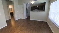This single-floor residence offers a clean, open-concept layout that seamlessly connects the main living area to the kitchen. Rich, dark hardwood-style flooring runs throughout, lending a warm and cohesive feel to the space. The living area is generously proportioned, providing ample room for furniture arrangement while maintaining an airy, uncluttered atmosphere. Overhead lighting illuminates the space, complementing the neutral-toned walls that create a versatile backdrop for a variety of interior styles. The kitchen is defined by a pass-through counter with stone-look countertops, offering both a visual separation and a convenient connection to the living area. Dark espresso-toned cabinetry provides substantial storage, while a pendant light adds a refined touch above the counter. Appliances, including a refrigerator, are integrated into the kitchen layout. A hallway leads to additional rooms, suggesting a practical and well-organized floor plan. Classic six-panel interior doors and white baseboards throughout add timeless architectural detail. Overall, the space presents a functional, well-appointed interior with consistent finishes that create a polished and inviting environment.