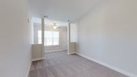 This single-floor, one-bedroom, one-bathroom residence is part of Phase II of the Autumn community and includes a desirable sunroom addition. The living area is finished with soft, neutral-toned carpeting and features vaulted ceilings that lend an open, airy feel to the space. Architectural column details frame the transition between the living and adjacent areas, adding a touch of structural character. A ceiling fan provides both comfort and subtle style overhead. Natural light flows generously through white-slatted blinds on double windows, brightening the interior with a clean, welcoming ambiance. The walls are painted in a warm, neutral tone throughout, creating a cohesive and calming atmosphere. The entryway, visible from the main living space, offers a classic paneled door and easy access to the rest of the home. The inclusion of a sunroom extends the livable space further, providing an ideal spot for relaxation or additional versatility. Ceiling vents are well-positioned for efficient climate control. Overall, the residence presents a tidy, move-in-ready environment with thoughtful design details suited for comfortable everyday living.
