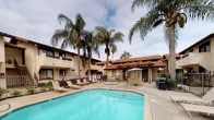 This residential community showcases a charming Spanish Colonial architectural style, characterized by warm stucco exteriors, rich terracotta tile roofing, and wood-accented balconies that lend the property a timeless, Southwest-inspired aesthetic. The multi-story buildings are arranged around a central outdoor amenity area that serves as the heart of the community.

The centerpiece of the outdoor space is a generously sized swimming pool with clear, inviting turquoise water, complemented by a separate spa/hot tub nearby. The expansive pool deck is finished in neutral concrete and is furnished with multiple lounge chairs, providing ample space for relaxation and sunbathing. A covered pergola area offers shaded seating with table and chairs, along with access to a communal barbecue grill — ideal for outdoor gatherings.

Tall, mature palm trees are planted throughout the courtyard, enhancing the resort-like ambiance and providing natural shade. Lush, manicured landscaping with flowering shrubs and greenery borders the pool area and surrounding walkways, adding color and vibrancy to the outdoor environment. Wrought iron fencing encloses the pool area for safety and privacy. The overall atmosphere is relaxed and sun-drenched, reflecting a comfortable California-style outdoor lifestyle.