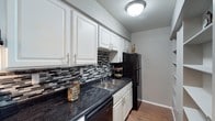This single-floor space showcases a thoughtfully designed kitchen that balances classic and contemporary elements. The kitchen is outfitted with crisp white cabinetry featuring brushed nickel hardware, providing ample upper and lower storage. Dark granite-style countertops offer a striking contrast against the white cabinets, while a mosaic tile backsplash in shades of black, white, and gray adds a dynamic visual element to the space. A double-basin stainless steel sink sits beneath the cabinetry, complemented by black appliances, including a refrigerator and dishwasher, that maintain a cohesive color palette throughout. Warm hardwood-style flooring runs underfoot, adding texture and warmth to the overall design. Overhead, a flush-mount ceiling light provides functional illumination. The kitchen's galley-style layout makes efficient use of space, with counters and cabinets lining one wall in a streamlined configuration. The neutral gray wall tones tie together the darker and lighter elements, resulting in a polished, modern aesthetic well-suited for everyday cooking and entertaining.