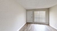 This single-story unit presents a clean, neutral aesthetic throughout, with consistent light-toned walls, white trim, and textured ceilings that unify the living spaces. The main living area features wood-look plank flooring and receives natural light through a large window fitted with horizontal blinds, positioned adjacent to the front entry door. The open-concept layout connects the living area to the kitchen via a peninsula counter with a light stone-style surface, providing both workspace and a casual breakfast bar. The kitchen is efficiently designed in a galley configuration, outfitted with stainless steel appliances including a refrigerator, range, and over-the-range microwave, complemented by gray cabinetry and continued wood-look flooring. The bedroom is carpeted in a neutral beige tone and is equipped with a ceiling fan with light fixture, as well as a window offering views of surrounding greenery, allowing natural light to filter through. The bathroom features wood-look tile flooring, a white vanity with a marble-style countertop, a large mirror, and a full tub and shower combination with white tile surround. The overall ambiance is bright, clean, and move-in ready.