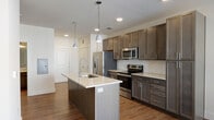 This contemporary single-floor residence is designed with a clean, modern aesthetic and thoughtful layout throughout. At the heart of the home is an open-concept kitchen and living area, where rich dark-toned cabinetry contrasts beautifully with light granite countertops. A central kitchen island with an undermount sink and pendant lighting serves as both a functional workspace and a natural gathering point. The kitchen is fully equipped with stainless steel appliances, including a French door refrigerator, electric range, and built-in microwave, all set against a classic white subway tile backsplash. Warm hardwood-style flooring extends throughout the main living areas, enhancing the sense of openness, while large windows allow natural light to flow in and offer views of the surrounding exterior. The bedroom features plush carpeting, a ceiling fan, and a bright window with blinds, creating a comfortable retreat. A generously sized walk-in closet with built-in rod storage provides ample organization space. The bathroom is finished with a granite-topped vanity, dark shaker-style cabinetry, a large framed mirror, and stylish industrial-inspired lighting, completing the modern and cohesive design of this well-appointed residence.