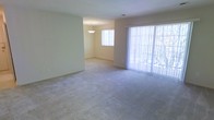 This single-floor residence offers a clean, functional layout designed to maximize comfort and usability. The open-plan living and dining area serves as the heart of the home, seamlessly connecting both spaces to create a generous, flowing environment ideal for everyday living and entertaining. Soft, neutral-toned carpeting extends across the floor, adding warmth and a sense of continuity throughout the space. The walls are finished in a light, creamy hue that enhances the overall brightness and contributes to an airy, welcoming ambiance. Natural light pours in through a large sliding glass door dressed with sheer vertical blinds, offering views of the outdoors while allowing flexible light control. A secondary window further brightens the dining area, which is anchored by a simple pendant light fixture that adds a touch of understated elegance. The transition between the living and dining zones is defined by a subtle architectural soffit, providing gentle spatial definition without compromising the open feel. The overall design is modest and move-in ready, presenting a neutral canvas that adapts easily to a variety of interior styles and furnishing preferences.