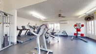 This single-floor fitness center offers a comprehensive workout environment designed to support a wide range of exercise routines. The space is organized into distinct functional zones, each catering to different training needs. The cardio area is outfitted with multiple treadmills, elliptical machines, and a stationary bike, all positioned near windows that allow natural light to filter in and provide views of the surrounding exterior. Overhead fluorescent lighting and ceiling fans ensure the space remains well-lit and comfortable throughout. The strength training section features a robust power rack with a full set of weight plates and barbells, complemented by a cable cross machine ideal for a variety of resistance exercises. A dedicated functional training area offers TRX suspension straps mounted to an overhead rig, a heavy tire for functional strength work, a rowing machine, a boxing station, and a yoga mat for stretching or floor exercises. The walls are finished in a clean, neutral tone with a textured ceiling, contributing to the space's practical and focused atmosphere. Multiple windows throughout provide ample daylight and a sense of openness, while a wall-mounted air conditioning unit supports climate control. The durable, low-maintenance flooring ties the entire space together, making this fitness center a well-rounded facility suited for diverse training goals.