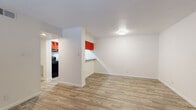 This single-story unit offers a functional and straightforward layout across several distinct areas. The living space near the front entry features durable wood-style plank flooring and a window that allows natural light to filter in, creating a bright and welcoming atmosphere. A flush-mount ceiling light and a visible HVAC vent contribute to the room's practical design.

The kitchen is a standout area, distinguished by bold red upper cabinetry that contrasts sharply with the dark lower cabinets and black appliances, including an electric range with overhead ventilation. Light-toned countertops provide workspace, and a pass-through counter connects the kitchen to an adjacent dining or flex area, which also features wood-style flooring.

The bedroom is carpeted for comfort and includes a ceiling fan with integrated lighting. Two doorways lead from the room — one to a walk-in closet equipped with shelving, and the other to additional areas of the unit. The bathroom is clean and utilitarian, offering a full tub and shower combination with white tile surround, a grab bar, and wood-style flooring. Overall, the unit presents a clean, move-in-ready environment with a cohesive, no-frills design.