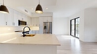 Located at 3420 Tamiami Trail E in Naples, Florida, this single-floor residence showcases a clean, contemporary design with a cohesive neutral palette and thoughtfully selected finishes throughout. Light wood-look flooring runs continuously across the entire space, enhancing the sense of openness and flow between rooms.

The kitchen serves as a central highlight, featuring an expansive quartz-topped island with an undermount sink and a matte black gooseneck faucet. White shaker-style cabinetry with black hardware extends to the ceiling, maximizing storage, while stainless steel appliances and a black dishwasher complete the modern aesthetic. Pendant lighting with dark metal fixtures adds a refined contrast above the island and perimeter counters.

Living areas benefit from abundant natural light delivered through oversized black-framed grid windows, which also frame views of surrounding greenery. A ceiling fan with a black finish provides both function and style. A black-framed glass door opens directly to a private outdoor deck, strengthening the indoor-outdoor connection.

Bedrooms are bright and airy, each equipped with large black-framed windows overlooking lush treetop views. Bathrooms are appointed with dual-sink floating vanities, white quartz countertops, black-framed mirrors, globe lighting, and neutral tile surrounds, delivering a polished, spa-like atmosphere.