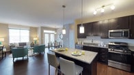 This single-floor residence showcases a thoughtfully designed open-concept layout that effortlessly connects the kitchen, dining, and living areas into one cohesive and inviting space. The kitchen is a standout feature, appointed with rich dark cabinetry, granite countertops, and a spacious central island with bar seating — ideal for casual dining or entertaining. Elegant pendant lighting hangs above the island, adding a warm, contemporary touch, while a subway tile backsplash and stainless steel appliances complete the polished look. The adjacent dining area benefits from natural light streaming through a set of classic French doors, creating a bright and airy atmosphere. The living area flows naturally from the dining space, offering a comfortable and relaxed setting. Throughout the space, dark hardwood flooring provides a strong visual anchor, beautifully contrasting with the neutral-toned walls. The overall ambiance is modern yet welcoming, with a carefully balanced mix of dark and light finishes that lend the space both sophistication and warmth. The open floor plan maximizes the sense of space, making the residence feel expansive and well-connected throughout.
