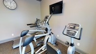 This single-floor fitness center is thoughtfully outfitted to meet a variety of cardio training needs. The space is anchored by a selection of modern cardio equipment, including elliptical trainers and an upright stationary bike, all featuring integrated digital consoles for tracking workout metrics. A wall-mounted flat-screen television is positioned at an ideal viewing height, allowing users to stay entertained or informed during their sessions. The room features neutral-toned walls that contribute to a clean, focused atmosphere, while the speckled carpet flooring adds a practical, durable surface underfoot. The layout is efficiently organized to maximize the use of the available floor space while maintaining comfortable access to each piece of equipment. The overall ambiance is functional and purposeful, providing a dedicated environment suited for consistent fitness routines. The space is well-suited for cardiovascular exercise, making it a convenient and practical amenity for its users.