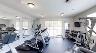 This fitness center is designed to provide a functional and motivating workout environment. The space is outfitted with a variety of cardio equipment, including elliptical trainers and treadmills, arranged to make efficient use of the floor area. A dedicated free weights section features a fully stocked dumbbell rack, catering to strength training needs. Large wall-mounted mirrors run along one side of the room, enhancing the sense of space and allowing users to monitor their form during exercise. The flooring is a dark, durable gym-grade material suited for high-traffic athletic use. Natural light floods the space through multiple large windows fitted with neutral roller shades, offering views of an outdoor balcony area and the surrounding environment. Ceiling-mounted flush lights provide consistent overhead illumination throughout. The walls are finished in a clean, bright white, contributing to an open and energizing atmosphere. A wall-mounted flat-screen television is also present, adding entertainment for longer workout sessions. The overall layout is thoughtfully organized to balance cardio, strength, and open floor space, creating a comprehensive and accessible fitness facility.
