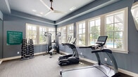 Located on Denali Drive in Mountain Park, GA, this fitness center is a thoughtfully designed workout space that offers a comprehensive suite of exercise equipment in a bright, welcoming environment. The room is finished with cool slate-blue walls, white trim, and neutral carpet flooring, creating a clean and focused atmosphere conducive to exercise.

Large, multi-pane windows line the walls, flooding the space with natural light and offering direct views of an outdoor pool and lush landscaping beyond. A ceiling fan with recessed lighting overhead ensures comfortable air circulation throughout workouts.

The fitness center is fully equipped with TRUE brand cardio machines, including treadmills and an elliptical, as well as a multi-station cable and strength training machine paired with an adjustable weight bench. Two sets of dumbbell racks hold a comprehensive range of free weights, catering to users of all fitness levels. Full-length wall mirrors expand the sense of space and allow for proper form checks during exercise.

Wall-mounted flat-screen televisions provide entertainment, while glass-paneled double doors open to an adjacent area, ensuring easy access. Fitness center rules are clearly posted, reflecting a well-managed and community-oriented facility. This space delivers a functional and well-appointed fitness experience.
