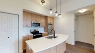 This single-floor residence showcases a thoughtfully designed open-concept layout with a strong emphasis on modern finishes and functional living. The kitchen serves as the heart of the space, featuring warm natural wood cabinetry that provides ample storage, complemented by a classic white subway tile backsplash that adds a clean, timeless aesthetic. Stainless steel appliances, including a French door refrigerator, electric range, and over-the-range microwave, outfit the kitchen with everything needed for everyday cooking. A standout feature is the generously sized curved kitchen island, finished with a crisp white quartz countertop and an undermount stainless steel sink with a matte black faucet, creating an elegant focal point ideal for both meal preparation and casual dining. Durable wood-look flooring flows throughout, grounding the space with a warm, cohesive tone. Lighting is a key design element, with a combination of a contemporary ceiling fixture and a trio of industrial-style mason jar pendant lights suspended above the island, adding character and charm. The open layout and light neutral wall tones contribute to an airy, welcoming ambiance throughout the space.