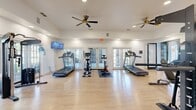 Located at 2350 Houston Lake Rd in Perry, Georgia, this fitness center offers a thoughtfully designed workout space spread across a single floor. The facility is organized into two connected areas, providing a comfortable and functional environment for a full range of exercise activities.

The main cardio area is bright and airy, featuring multiple treadmills, an elliptical trainer, and a stationary spin bike. Large French doors line the far wall, flooding the space with natural light and offering views of the outdoors. Two ceiling fans with gold-toned fixtures provide steady airflow throughout, while a wall-mounted flat-screen television adds entertainment for cardio sessions. Recessed lighting and white walls contribute to the clean, open atmosphere.

The adjoining strength and functional training area is equally well-appointed, housing a multi-station cable machine, a dedicated dumbbell rack stocked with a range of weights, kettlebells, medicine balls, and resistance training accessories. A large mirror runs along one wall to assist with form, and a ceiling fan keeps the area comfortable during intense workouts.

Throughout both sections, light hardwood-style flooring provides a clean, modern look while remaining durable underfoot. The overall design is practical yet inviting, offering residents a comprehensive fitness facility that supports both cardiovascular and strength training goals.