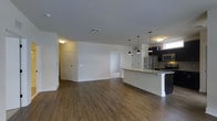 This single-floor space showcases a contemporary open-concept design that seamlessly integrates the living area and kitchen into one cohesive layout. The flooring throughout is a rich, dark hardwood-style material that adds warmth and contrast against the crisp white walls and light grey ceiling.

The kitchen is a standout feature, equipped with stainless steel appliances including a refrigerator and microwave. Dark cabinetry provides a bold visual contrast to the light-toned granite or stone countertops. A peninsula-style island with an undermount sink extends into the living space, offering additional prep space and a natural gathering point. Three sleek pendant lights hang above the island, providing both task lighting and a modern aesthetic touch.

The open living area is generously proportioned, offering flexibility for various furniture arrangements. Recessed ceiling vents ensure efficient climate control throughout. Multiple doorways visible from the main space suggest additional rooms beyond, while a window near the kitchen allows natural light to filter in. The overall ambiance is clean, modern, and move-in ready.