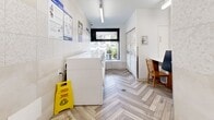 Located at 4222 Bathurst Street in Toronto, this self-contained laundry facility offers a clean, well-appointed space designed for practical, everyday use. The room is finished with eye-catching herringbone-patterned wood-look tile flooring that runs throughout, lending a modern yet warm aesthetic to the utilitarian space. Walls are clad in large-format cream and beige tiles, with upper sections featuring a subtle hexagonal mosaic pattern that adds a refined textural detail. A row of top-load washing machines lines one side of the room, while a bank of white storage lockers occupies the opposite wall, providing convenient secure storage for users. A folding or work surface with seating is available near the entrance, offering a practical spot for managing laundry tasks. Bright overhead fluorescent lighting ensures the space is well-lit throughout, complemented by a large street-facing window that brings in natural light and a view of the surrounding neighbourhood. Entry is through a glass-panelled door equipped with a door closer, ensuring controlled access. A fire extinguisher is mounted near the entrance, reflecting attention to safety standards. The overall ambiance is tidy, functional, and thoughtfully finished, making this a comfortable shared amenity for building residents.