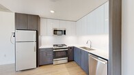 This thoughtfully designed urban space showcases a striking blend of industrial and modern aesthetics across a single, well-appointed floor. Exposed concrete ceilings and structural columns provide a bold architectural contrast against crisp white walls and light-toned hardwood flooring that flows throughout the main living areas. The open-concept living and kitchen area is anchored by dramatic floor-to-ceiling windows that wrap around the corner, flooding the space with natural light and offering panoramic views of the city skyline, a bridge, and a nearby waterway.

The kitchen is outfitted with a full suite of stainless steel appliances, including a gas range, built-in microwave, dishwasher, and refrigerator. Two-toned cabinetry — featuring sleek white upper cabinets and textured dark lower cabinets — frames a clean white countertop and undermount sink, creating a functional yet visually refined workspace.

The bedroom benefits from the same expansive window system, with carpet underfoot and abundant natural light. The bathroom is finished with large-format tile flooring, a wide vanity with dark cabinetry and white countertop, and a full-width mirror. A dedicated closet provides additional storage. The overall ambiance is clean, contemporary, and urban.
