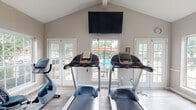 Located at 1500 Brentridge Dr in Nashville, Tennessee, this fitness center is a well-appointed, single-story facility designed to support an active lifestyle. The room features a striking vaulted ceiling that lends a sense of openness and airiness to the workout space. Light wood-style flooring runs throughout, complementing the neutral gray walls and crisp white trim.

The fitness center is equipped with multiple treadmills, a recumbent bike, an elliptical machine, a stair climber, a weight bench, and a rack of free weights, offering a well-rounded selection of cardio and strength training options. A wall-mounted flat-screen television provides entertainment during workouts.

Natural light floods the space through an abundance of large windows and sets of French doors that open toward an outdoor pool area, offering pleasant views of the pool and surrounding patio with its colorful umbrellas. A water cooler station is conveniently positioned near the equipment, and a paper towel dispenser is mounted on the wall for easy access.

A vibrant, colorful piece of wall art adds a lively touch to the otherwise clean and functional aesthetic. Posted fitness center rules and clearly marked exit signage reflect a commitment to safety and community standards. The space is bright, welcoming, and thoughtfully laid out for efficient use.