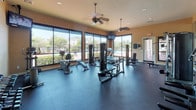 This single-floor fitness center is designed to support a full range of workout routines, offering a thoughtfully organized and generously proportioned exercise space. The layout accommodates both strength training and cardiovascular equipment, with ample room to move freely between stations.

Large windows along one wall flood the interior with natural light while providing views of the surrounding outdoor landscape, including lush greenery and neighboring structures. Ceiling fans are mounted overhead, contributing to comfortable air circulation throughout the space. Recessed lighting supplements natural light, ensuring consistent illumination across the room.

The strength training area includes a multi-functional cable machine, a rack of dumbbells in graduated weights, medicine balls on a dedicated stand, a weight bench, and a body weight scale. The cardio section is well-appointed with treadmills, elliptical trainers, a stationary bike, and a rowing machine positioned to take advantage of the window views.

Wall-to-wall mirrors on one side visually expand the space and assist with form during exercise. The durable rubber flooring throughout provides cushioning and safety. A wall-mounted television is also present, adding an entertainment option during workouts. The warm, neutral-toned walls and natural light create an energizing yet comfortable atmosphere.