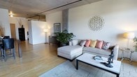 This single-floor space showcases a compelling blend of industrial and contemporary design. The open-plan living area is defined by its exposed concrete structural columns and visible overhead piping, hallmarks of classic loft-style architecture that lend the space a distinctive urban character. Warm hardwood flooring runs throughout, providing a natural contrast to the raw industrial elements above.

The living area is generously proportioned, offering an open and airy feel with high ceilings that amplify the sense of space. A large sectional sofa with a chaise lounge serves as the centerpiece, accented by a collection of colorful decorative pillows in pink, coral, and yellow tones. A sleek rectangular coffee table anchors the seating area, which sits atop a neutral area rug. Decorative wall art — including a striking metallic sunburst piece — adds personality and warmth to the neutral-toned walls.

Natural light fills the space, complemented by ambient floor lamps that create a welcoming atmosphere. Lush indoor greenery adds a refreshing organic element, softening the industrial aesthetic. The open layout allows for a seamless flow between the living area and the additional open space beyond, making this a versatile and inviting environment well-suited for both relaxed living and entertaining.