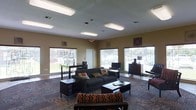 This single-floor space is designed as a comfortable and functional communal gathering area. The open-plan layout provides a generous sense of space, anchored by a central seating arrangement that includes a dark upholstered sofa accented with colorful throw pillows and a coordinating coffee table. Additional seating options, including armchairs and accent pieces, are thoughtfully arranged throughout the room to encourage conversation and relaxation.

Natural light flows in abundantly through multiple large windows fitted with horizontal blinds, offering views of the surrounding greenery outside. The warm, neutral-toned walls are complemented by flush-mounted ceiling light fixtures that ensure the space remains bright and well-lit throughout the day. Decorative wall art adds personality and visual interest to the room.

Along the perimeter, a wooden dresser with a lamp serves as a functional accent, while a desk area near the windows provides a dedicated workspace. The flooring features a patterned carpet that adds texture and warmth underfoot. The overall ambiance is casual yet purposeful, making this space well-suited for both social gatherings and quiet individual use. Its straightforward, practical design emphasizes accessibility and comfort for all who use it.