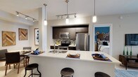 This single-floor space showcases a thoughtfully designed open-concept layout that connects the kitchen, dining, and sleeping areas in a cohesive and functional arrangement. The kitchen is a standout feature, outfitted with stainless steel appliances including a refrigerator, microwave, and range, complemented by dark espresso cabinetry that extends to the ceiling for ample storage. A decorative mosaic tile backsplash adds texture and visual interest behind the cooking area. The light-toned countertops provide a striking contrast to the dark cabinetry, and the peninsula-style breakfast bar extends the counter space while accommodating bar stools for casual dining or entertaining. Pendant lighting in varying styles hangs above the bar, adding a modern, layered lighting scheme alongside recessed track lighting overhead. A built-in sink is integrated into the peninsula for added convenience. Beyond the kitchen, a glimpse into the bedroom reveals a serene space accented by a bold teal feature wall adorned with decorative circular art pieces. The neutral white walls throughout keep the space feeling bright and open, while cohesive design elements tie each area together seamlessly.