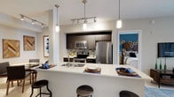 This single-floor space showcases a thoughtfully designed open-concept layout that connects the kitchen, dining, and sleeping areas in a cohesive and functional arrangement. The kitchen is a standout feature, outfitted with stainless steel appliances including a refrigerator, microwave, and range, complemented by dark espresso cabinetry that extends to the ceiling for ample storage. A decorative mosaic tile backsplash adds texture and visual interest behind the cooking area. The light-toned countertops provide a striking contrast to the dark cabinetry, and the peninsula-style breakfast bar extends the counter space while accommodating bar stools for casual dining or entertaining. Pendant lighting in varying styles hangs above the bar, adding a modern, layered lighting scheme alongside recessed track lighting overhead. A built-in sink is integrated into the peninsula for added convenience. Beyond the kitchen, a glimpse into the bedroom reveals a serene space accented by a bold teal feature wall adorned with decorative circular art pieces. The neutral white walls throughout keep the space feeling bright and open, while cohesive design elements tie each area together seamlessly.
