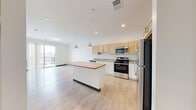 This well-designed 1-bedroom, 1-bathroom unit offers a thoughtful layout across a single floor, maximizing functionality within a compact footprint. The open-concept living and kitchen area serves as the heart of the unit, featuring light wood laminate flooring, crisp white walls, and recessed lighting complemented by stylish pendant lights above a central kitchen island. The kitchen is equipped with stainless steel appliances—including a refrigerator, gas range, microwave, and dishwasher—alongside warm-toned wood cabinetry and ample counter space. A sliding door leads to a balcony, allowing natural light to flow into the living area. The bedroom offers a cozy, carpeted retreat with two large windows that frame outdoor views, providing generous natural light. The bedroom is served by a dedicated walk-in closet featuring wire shelving that runs the full width of the space, offering practical storage. The bathroom includes a clean, all-white walk-in shower. A hallway connects the living spaces to the private quarters, contributing to the unit's efficient flow. The overall aesthetic is contemporary and clean, with a neutral palette throughout that creates a bright, inviting atmosphere.