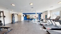 Located at 196 Mansfield Avenue in Norton, MA, this fitness center offers a comprehensive exercise facility designed to support a wide range of workout routines. The single-floor space is thoughtfully laid out to maximize functionality and ease of movement throughout.

The interior features a clean, modern aesthetic with crisp white walls accented by bold blue horizontal stripe detailing, creating an energizing atmosphere. Durable speckled flooring runs throughout, providing a practical and polished surface suited for high-traffic use. Recessed and flush-mount ceiling fixtures provide consistent overhead lighting, complemented by generous natural light streaming in through a row of large windows along one wall, offering views of the surrounding outdoor area.

The cardio zone is well-appointed with multiple treadmills, elliptical machines, a rowing machine, and a stationary bike. A large wall-mounted mirror expands the sense of space and allows users to monitor their form during workouts. A wall-mounted flat-screen TV adds to the functionality of the cardio area.

The strength training section includes a power rack with weight plates, a cable machine, a dumbbell rack, and a weight bench, providing a full complement of resistance training options. Glass double doors connect the space to the broader building, and sanitation stations are conveniently positioned near the entrance. Colorful wall art adds a vibrant finishing touch to this well-rounded fitness facility.