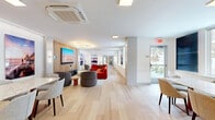 Located at 22 Hubbard Road in Wilton, Connecticut, this single-floor communal space offers a thoughtfully designed environment that seamlessly blends a living lounge with a dedicated dining area. The open layout is anchored by light-toned hardwood flooring that flows continuously throughout, lending the space a clean, cohesive aesthetic.

The living area serves as a vibrant social hub, highlighted by a bold red sectional sofa, rounded upholstered accent chairs, and a striking circular LED chandelier that draws the eye upward. A warm wood-paneled feature wall frames a large flat-screen television, creating an inviting media focal point. Curated large-format landscape photography is displayed throughout, adding color and a sense of dynamic artistic character to the white walls.

The dining area offers a spacious granite-topped table surrounded by upholstered side chairs, with natural light streaming in through generous windows that overlook the surrounding neighborhood. A built-in wood shelving unit with decorative objects adds warmth and a refined finishing touch to the dining zone.

Additional seating nooks with rounded lounge chairs and small accent tables are interspersed throughout, providing flexible spaces for conversation or quiet relaxation. The overall ambiance is contemporary and welcoming, combining modern design sensibilities with functional, community-oriented living.