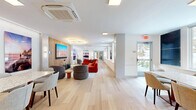 Located at 22 Hubbard Road in Wilton, Connecticut, this single-floor communal space offers a thoughtfully designed environment that seamlessly blends a living lounge with a dedicated dining area. The open layout is anchored by light-toned hardwood flooring that flows continuously throughout, lending the space a clean, cohesive aesthetic.

The living area serves as a vibrant social hub, highlighted by a bold red sectional sofa, rounded upholstered accent chairs, and a striking circular LED chandelier that draws the eye upward. A warm wood-paneled feature wall frames a large flat-screen television, creating an inviting media focal point. Curated large-format landscape photography is displayed throughout, adding color and a sense of dynamic artistic character to the white walls.

The dining area offers a spacious granite-topped table surrounded by upholstered side chairs, with natural light streaming in through generous windows that overlook the surrounding neighborhood. A built-in wood shelving unit with decorative objects adds warmth and a refined finishing touch to the dining zone.

Additional seating nooks with rounded lounge chairs and small accent tables are interspersed throughout, providing flexible spaces for conversation or quiet relaxation. The overall ambiance is contemporary and welcoming, combining modern design sensibilities with functional, community-oriented living.
