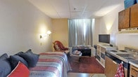This single-floor space is designed as a compact, self-contained room that efficiently combines sleeping, seating, and entertainment areas within one open layout. The room features a drop tile ceiling and textured walls painted in warm beige tones, contributing to a cozy, understated ambiance.

The sleeping area is anchored by a full-sized bed dressed in a patterned, multicolored quilt, accompanied by a wall-mounted swing-arm lamp that provides focused bedside lighting. Adjacent to the bed, a cushioned armchair draped with a throw blanket offers a comfortable spot for relaxing.

Natural light filters into the room through a window dressed with floor-length gray curtains, which can be drawn for privacy. A low, dark-toned media console sits beneath the window, topped with a flat-screen television and complemented by a small coffee table nearby. To the right, a light-colored storage dresser with mixed drawer fronts provides additional organization and storage.

Dark hardwood-style flooring runs throughout the space, adding warmth and grounding the overall aesthetic. The room's layout maximizes functionality, making efficient use of its available footprint while maintaining a livable, practical environment.