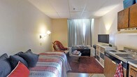This single-floor space is designed as a compact, self-contained room that efficiently combines sleeping, seating, and entertainment areas within one open layout. The room features a drop tile ceiling and textured walls painted in warm beige tones, contributing to a cozy, understated ambiance.

The sleeping area is anchored by a full-sized bed dressed in a patterned, multicolored quilt, accompanied by a wall-mounted swing-arm lamp that provides focused bedside lighting. Adjacent to the bed, a cushioned armchair draped with a throw blanket offers a comfortable spot for relaxing.

Natural light filters into the room through a window dressed with floor-length gray curtains, which can be drawn for privacy. A low, dark-toned media console sits beneath the window, topped with a flat-screen television and complemented by a small coffee table nearby. To the right, a light-colored storage dresser with mixed drawer fronts provides additional organization and storage.

Dark hardwood-style flooring runs throughout the space, adding warmth and grounding the overall aesthetic. The room's layout maximizes functionality, making efficient use of its available footprint while maintaining a livable, practical environment.