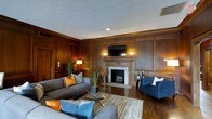 This distinguished single-floor space exudes classic elegance through its rich, floor-to-ceiling wood-paneled walls, which lend a sense of warmth and refined craftsmanship throughout. The living area serves as the heart of the space, anchored by a striking fireplace with a crisp white surround that provides a tasteful contrast against the deep wood tones. A flat-screen television is mounted prominently above the fireplace, flanked by wall sconces that cast a soft, ambient glow. The hardwood flooring complements the paneled walls, creating a cohesive and timeless aesthetic. Comfortable seating arrangements include a generous gray sofa adorned with orange and blue accent pillows, paired with a sleek blue tufted armchair, offering an inviting and stylish lounge setting. A glass-top coffee table with a sculptural metal base anchors the seating area, while potted greenery adds a touch of natural life to the interior. Recessed lighting and a textured ceiling complete the well-appointed look. Overall, the space balances traditional architectural detail with contemporary furnishings, resulting in an atmosphere that is both sophisticated and welcoming.