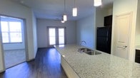 This single-story space showcases an open-concept layout that seamlessly connects the kitchen and living areas. The kitchen is a standout feature, highlighted by an expansive granite countertop island with an undermount stainless steel sink and modern pendant lighting hanging above. Black appliances, including a full-size refrigerator, complement the contemporary aesthetic. The dark hardwood-style flooring runs throughout, providing a rich contrast to the light-toned walls and countertops. The living area is illuminated by a ceiling fan with integrated lighting, contributing to both comfort and functionality. Natural light flows into the space through a set of French doors, which open to an outdoor area, enhancing the sense of openness. The neutral wall tones and clean lines throughout give the space a modern, move-in-ready feel. The thoughtful design of the open floor plan allows for flexible furniture arrangements and makes the overall space feel expansive and well-connected.