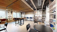 Located at 49 Fisk St in Jersey City, NJ, this single-floor collaborative lounge space offers a compact yet efficiently arranged environment designed for productivity and comfort. The most striking architectural feature is the large-scale woven rope ceiling installation, which adds warmth and acoustic texture while contrasting beautifully with the exposed industrial ceiling elements, including black structural beams and track lighting.

The space is organized into several distinct seating zones. A live-edge communal high-top table anchors the central area, surrounded by modern bar-height chairs in a mix of blue and dark tones — ideal for collaborative work or casual gatherings. Along the perimeter, built-in window-facing desks with individual task lighting and terracotta-hued chairs offer quieter, focused work spots, framed by large windows that flood the space with natural light and provide outward views of the surrounding neighborhood.

A built-in bench nook lines one wall, paired with small terrazzo-topped bistro tables and flanked by open wood shelving units displaying books, plants, and curated décor. Additional round tables and black chairs are distributed throughout, accommodating a variety of uses. Light cork flooring ties the warm wood tones together, creating a cohesive, inviting atmosphere throughout this well-appointed space.