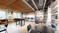 Located at 49 Fisk St in Jersey City, NJ, this single-floor collaborative lounge space offers a compact yet efficiently arranged environment designed for productivity and comfort. The most striking architectural feature is the large-scale woven rope ceiling installation, which adds warmth and acoustic texture while contrasting beautifully with the exposed industrial ceiling elements, including black structural beams and track lighting.

The space is organized into several distinct seating zones. A live-edge communal high-top table anchors the central area, surrounded by modern bar-height chairs in a mix of blue and dark tones — ideal for collaborative work or casual gatherings. Along the perimeter, built-in window-facing desks with individual task lighting and terracotta-hued chairs offer quieter, focused work spots, framed by large windows that flood the space with natural light and provide outward views of the surrounding neighborhood.

A built-in bench nook lines one wall, paired with small terrazzo-topped bistro tables and flanked by open wood shelving units displaying books, plants, and curated décor. Additional round tables and black chairs are distributed throughout, accommodating a variety of uses. Light cork flooring ties the warm wood tones together, creating a cohesive, inviting atmosphere throughout this well-appointed space.