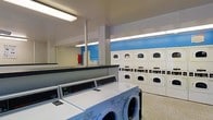 This single-floor laundry facility is purpose-built for commercial or communal use, offering a straightforward and functional layout designed to maximize efficiency. The main area houses a row of front-loading washing machines aligned along one side, providing ample washing capacity for high-volume use. Opposite the washers, stacked dryer units line the wall in a neat, organized configuration, making effective use of vertical space and accommodating multiple users simultaneously. The walls are painted in a clean white and blue color scheme, contributing to a bright and tidy atmosphere. Overhead, fluorescent lighting ensures the entire space is evenly and thoroughly illuminated. Structural columns are positioned throughout the room, adding to the industrial character of the facility. A service counter or folding area is also present, offering a practical surface for sorting and organizing laundry. The flooring is durable and easy to maintain, suited to the demands of a high-traffic environment. A fire extinguisher is visibly mounted on the wall, reflecting attention to safety. Overall, the space is well-organized, utilitarian, and designed with functionality and user convenience at the forefront.