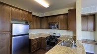 This single-floor space showcases a thoughtfully designed kitchen that combines functionality with a warm, inviting aesthetic. The kitchen is outfitted with rich, dark wood cabinetry that extends to the upper walls, providing generous storage throughout. Cabinet doors feature simple, classic hardware that complements the overall design. The countertops are finished in light granite with subtle veining, offering a pleasing contrast against the darker cabinetry below.

Stainless steel appliances include a refrigerator, a built-in microwave, and a gas range with multiple burners and an integrated oven, all contributing to a cohesive, modern look. A stainless steel sink with a contemporary faucet is conveniently positioned along the counter. The layout follows an efficient L-shaped configuration, maximizing workspace and ease of movement during food preparation.

Hardwood-style flooring grounds the space with a warm tone that ties together the cabinetry and countertop finishes. Overhead, flush-mounted lighting provides even illumination throughout the kitchen. A ceiling vent ensures proper ventilation, adding to the practicality of the space. The overall ambiance is clean, functional, and well-organized.