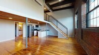 This two-story loft in Providence, Rhode Island, showcases a compelling blend of industrial heritage and modern updates. The main level is defined by rich, warm hardwood floors that extend throughout the open living area, anchored by exposed brick walls and large multi-pane windows that frame city views and flood the space with natural light. Structural steel columns and dark overhead beams reinforce the industrial character of the space.

A contemporary kitchen features white shaker cabinetry, gray stone countertops, and stainless steel appliances including a refrigerator, electric range, microwave, and dishwasher. A dedicated laundry closet with a stacked washer and dryer adds practical convenience. The main-level bathroom includes a white vanity with matching countertop and hardwood flooring beneath a wood-paneled ceiling.

A sleek open staircase with a black metal railing leads to the upper level, where exposed dark timber beams add dramatic character. The upper floor features carpeted bedroom space with a walk-in closet, offering a quieter retreat from the open lower level. The wood-paneled ceilings present throughout the space tie both levels together, creating a cohesive, loft-style aesthetic throughout.