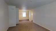 This single-story space presents a straightforward, functional layout with a clean and neutral aesthetic throughout. The interior features a combination of flooring types, with plush carpet covering the main living area and hardwood-style flooring visible in the adjoining room, offering a practical distinction between spaces.

The walls are painted in a soft, warm neutral tone that contributes to a bright and welcoming atmosphere. White baseboards run along the perimeter, providing a crisp contrast to the flooring and walls. The textured ceiling adds subtle visual interest while housing a centrally located HVAC vent, indicating a built-in climate control system.

The adjacent room, partially visible beyond an open doorway, benefits from natural light streaming through a window, enhancing the overall brightness of the space. A chandelier-style light fixture hangs in this area, serving as a modest decorative focal point. An additional doorway or closet opening is visible along the wall, suggesting practical storage or room access options.

Overall, the space maintains a clean, move-in-ready appearance with a simple, understated design suitable for a variety of uses and furnishing styles.