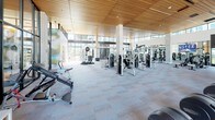 Located at 15400 NE 20th St in Bellevue, WA, this expansive fitness center is a thoughtfully designed, single-floor facility that offers a comprehensive workout experience. The space is defined by its striking architectural character, including warm wood-slat ceilings, floor-to-ceiling black-framed windows, and an abundance of natural light that fills the interior throughout the day.

The facility is organized into several distinct zones. The cardio area features a full row of treadmills, elliptical trainers, and rowing machines arranged to maximize space and airflow. Adjacent to this is a dedicated cycling studio with a large display screen and hardwood flooring, offering an immersive group fitness environment. A separate functional training zone is equipped with RAX multi-functional rig systems, TRX suspension trainers, medicine balls, battle ropes, a punching bag, and a variety of free weights and accessories. The strength training section houses an extensive selection of selectorized cable machines and weight benches.

Large glass partition walls divide the zones while maintaining visual connectivity across the facility. Mirrored walls further enhance the sense of openness. The overall ambiance is bright, clean, and professional, striking a balance between high-end design and practical athletic utility, making it a well-appointed destination for a wide range of fitness needs.