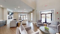This single-floor community space is designed with a contemporary aesthetic, combining functionality and comfort throughout its open layout. The main lounge area is generously proportioned, featuring soaring ceilings that enhance the sense of spaciousness. Neutral warm-toned walls are complemented by wood-look tile flooring laid in a herringbone pattern, creating a cohesive and inviting atmosphere. Large floor-to-ceiling windows and glass entry doors flood the interior with natural light while providing views of the surrounding exterior courtyard and buildings.

The lounge is furnished with multiple plush seating arrangements, including upholstered sofas and accent chairs in neutral tones, accented by natural wood side tables. A wall-mounted fireplace with a built-in television above serves as a focal point for one of the seating areas, while a beverage station with gray cabinetry offers added convenience nearby.

An adjacent kitchenette is well-equipped with gray shaker-style cabinetry, white quartz countertops, a stainless steel refrigerator, microwave, and double sink. A peninsula with bar-height seating accommodates casual dining or gathering. Additional cabinetry and prep space line the walls, making the area practical for events and everyday use. Recessed lighting throughout ensures the space remains bright and welcoming.