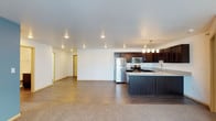 This single-floor residence offers a thoughtfully designed open-concept layout that seamlessly connects the kitchen and living areas into one cohesive, airy space. The kitchen is a standout feature, outfitted with rich dark wood cabinetry, granite-look countertops, and a full suite of stainless steel appliances including a refrigerator, microwave, and gas range. A peninsula with an undermount sink and pendant lighting above adds both functionality and a polished aesthetic. Recessed lighting throughout the ceiling ensures the space remains bright and welcoming. The living area flows naturally from the kitchen, featuring warm-toned carpet that contrasts with the hardwood-style flooring in the kitchen zone, creating a subtle yet effective delineation between spaces. Clean white walls and a smooth ceiling contribute to a fresh, neutral palette that offers versatility for any interior style. A hallway visible toward the rear of the space leads to additional rooms, suggesting a practical and well-organized floor plan. Overall, this residence combines modern finishes with an open, functional layout suited for comfortable everyday living.