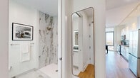 Located at 8601 Bergenline Avenue in North Bergen, NJ, this thoughtfully designed studio offers a modern, efficient layout that maximizes every square foot. The open-plan kitchen and living area serves as the heart of the space, featuring warm hardwood flooring, crisp white shaker cabinetry extending to the ceiling, and a full suite of stainless steel appliances including a French door refrigerator, electric range with over-the-range microwave, and dishwasher. A subtle tile backsplash and clean countertops complete the kitchen's polished aesthetic. The living area is anchored by a dramatic floor-to-ceiling window that floods the space with natural light and frames sweeping panoramic views of the surrounding landscape — a true standout feature. A hallway connects the living space to the bathroom and three closets, providing generous storage. The bathroom is finished to an elevated standard, showcasing large-format marble-look tile from floor to ceiling, a walk-in shower, and brushed gold fixtures that lend a luxurious touch. An in-unit laundry area adds everyday convenience. Throughout the space, a bright, neutral palette and consistent hardwood flooring create a cohesive, contemporary ambiance.